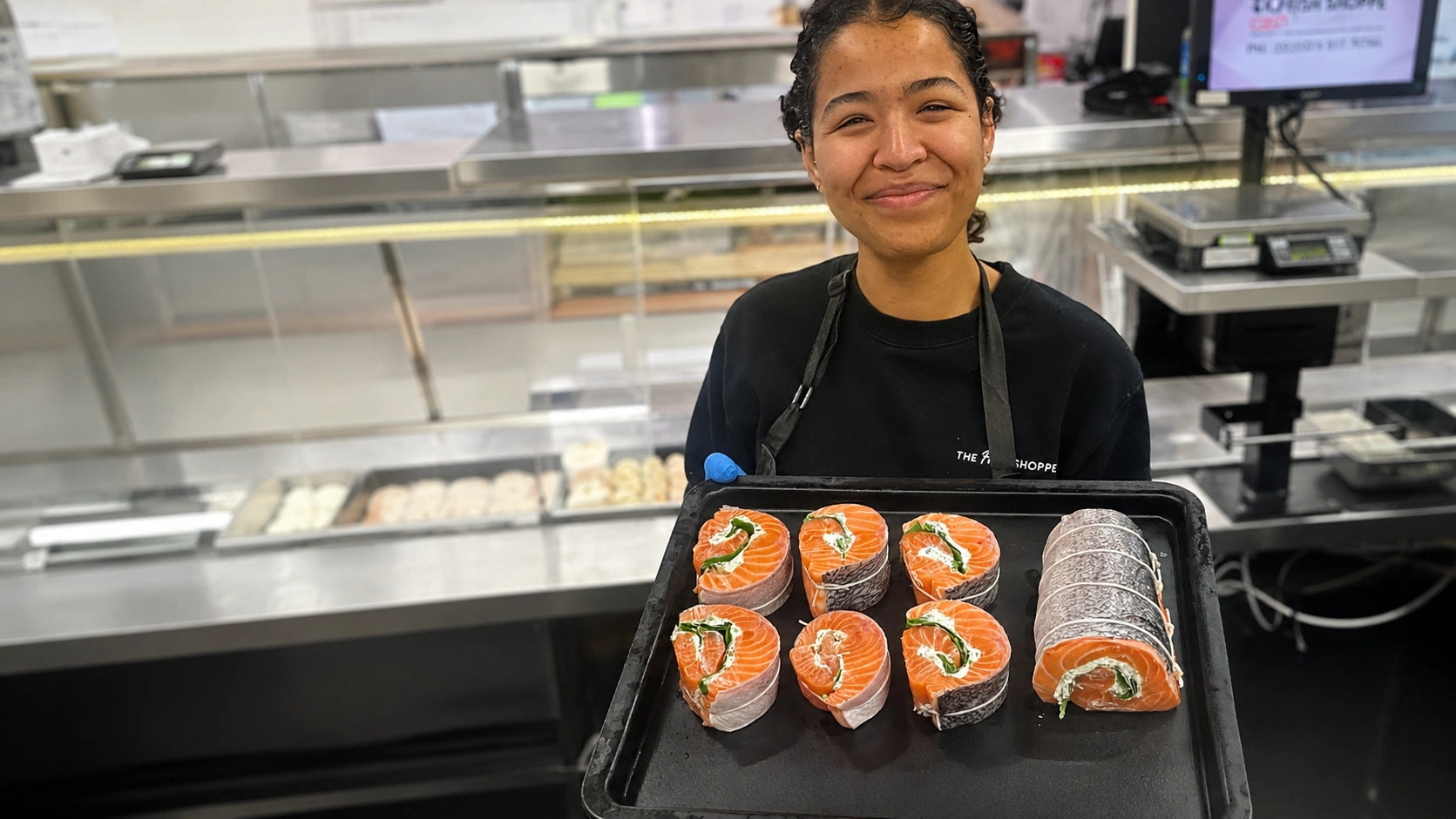 A smiling woman in a black uniform stands behind a counter holding a black tray with salmon sushi rolls wrapped in nori and green seaweed salad, with a commercial kitchen background.