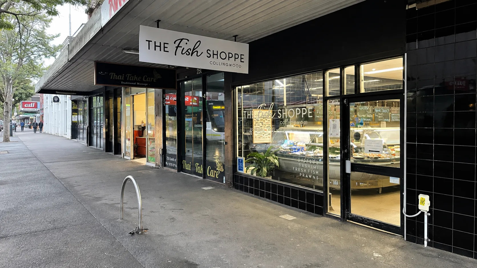 The Fish Shoppe storefront on a city sidewalk, with large glass windows, a white hanging sign, a black tiled facade, and a freestanding chalkboard sign on the sidewalk.