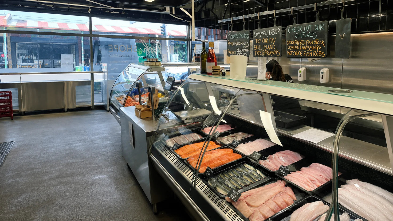 A seafood display case in a grocery store showing fresh fish, including pink fillets, silver whole fish, and orange salmon, with glass shelving and fluorescent lighting.