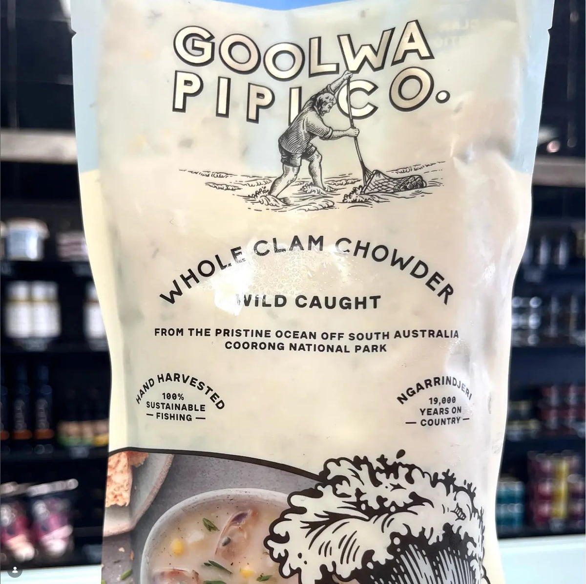 A stand-up pouch of Goolwa Pipi Co. Whole Clam Chowder Wild Caught, featuring a cream-colored label with black text and illustrations, set against a blurred dark retail background.