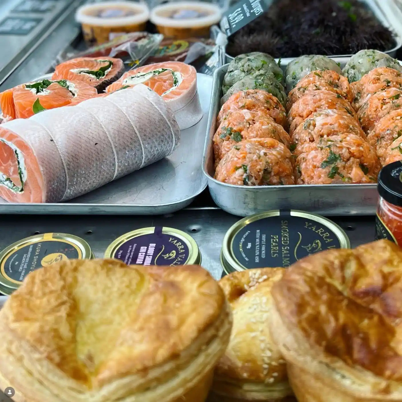A food display featuring salmon salmon pitta wraps, smoked salmon rolls wrapped in skin, and golden flaky pastries in the foreground, with small packaged items in the background.