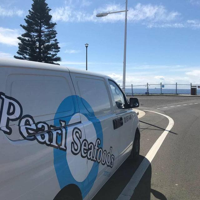 A white Pearl Seafoods van parked on an outdoor road with a blue sky, ocean, and trees in the background.