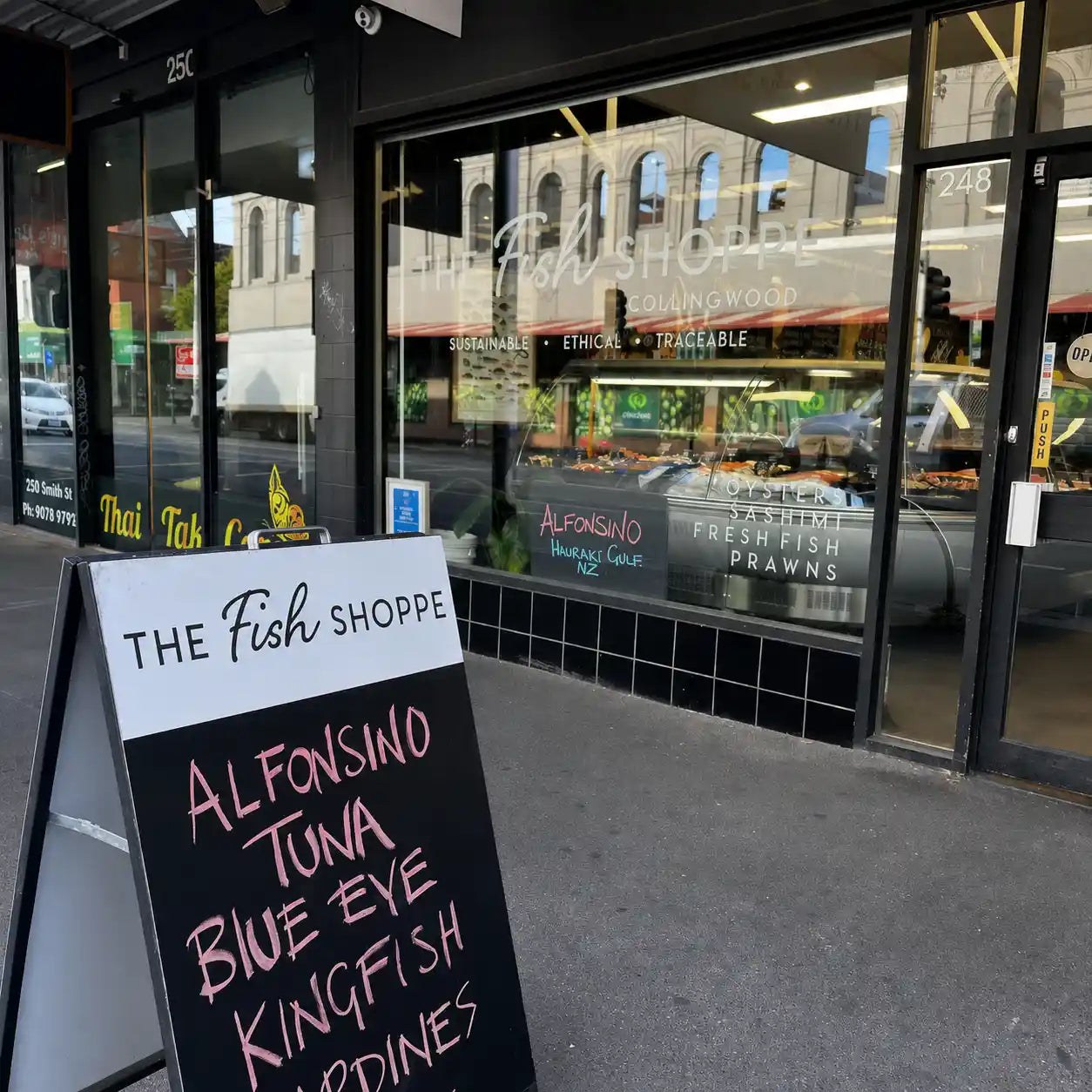 A storefront scene for The Fish Shoppe with large glass windows showing a seafood display case inside, and a black sidewalk A-frame sign in front reading Alfonsino Tuna Blue Eye Kingfish and others.
