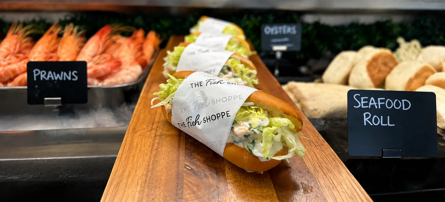A seafood roll sandwich from The Fish & Food Shoppe, wrapped in white paper, displayed on a wooden board in a restaurant setting with prawns and oysters in the background.