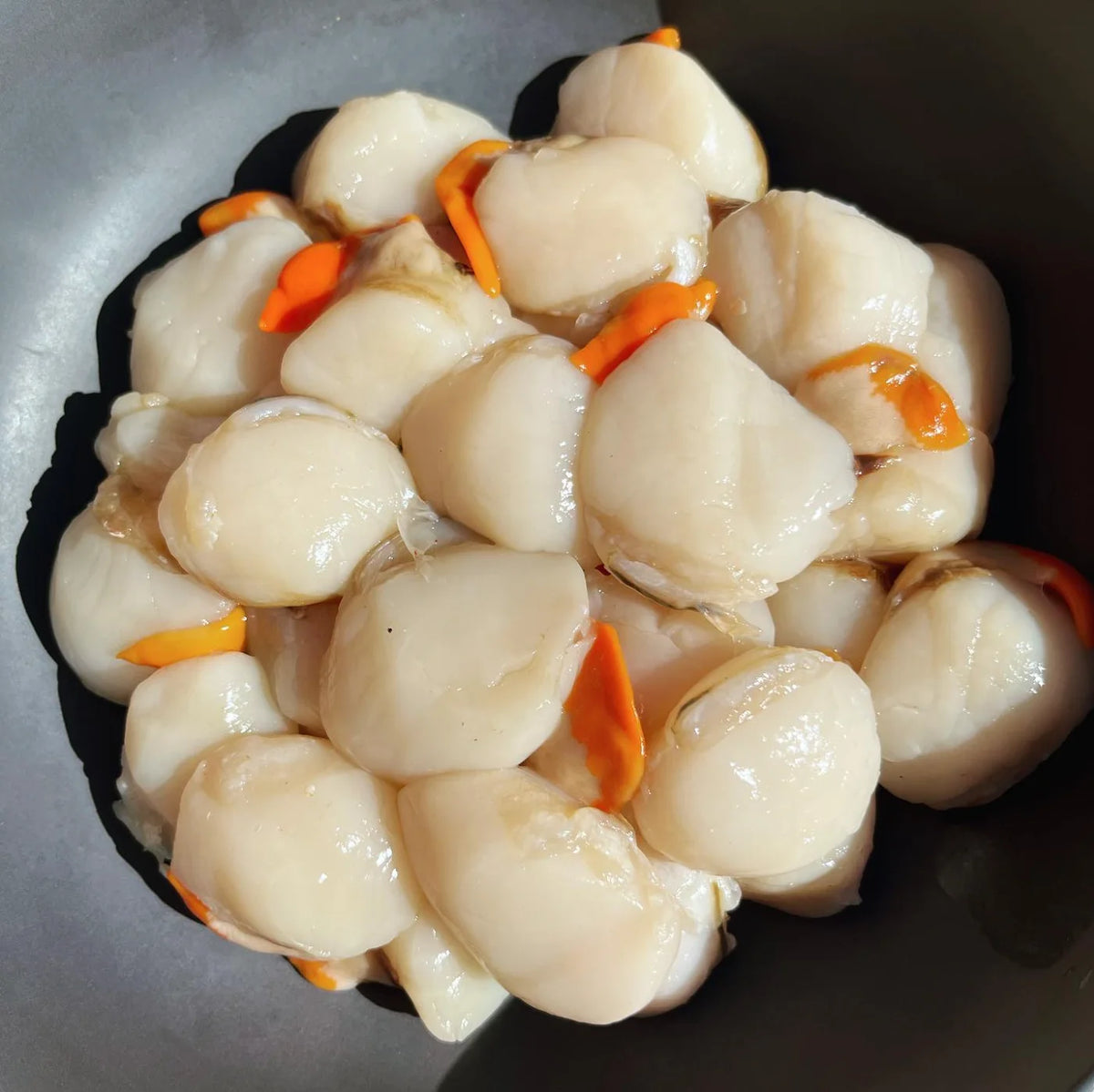 Raw scallop shellfish pieces cooked in a dark pan with small orange carrot slices visible among them.