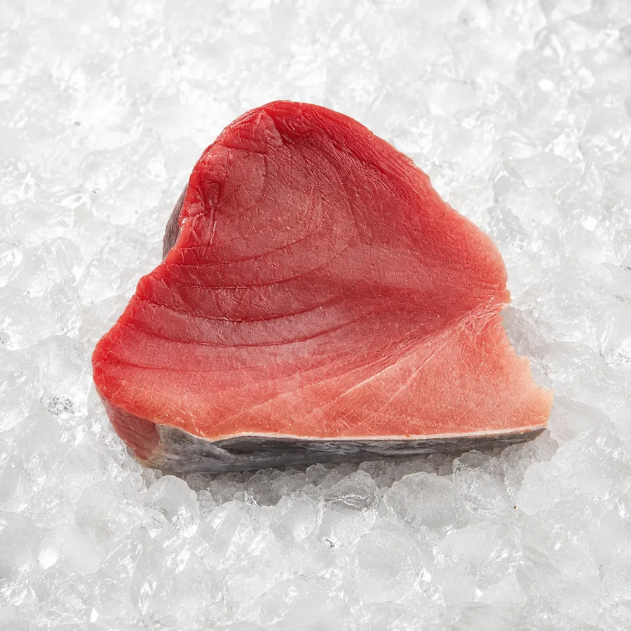 A thick slice of raw tuna steak with a dark skin layer, resting on a bed of crushed ice.