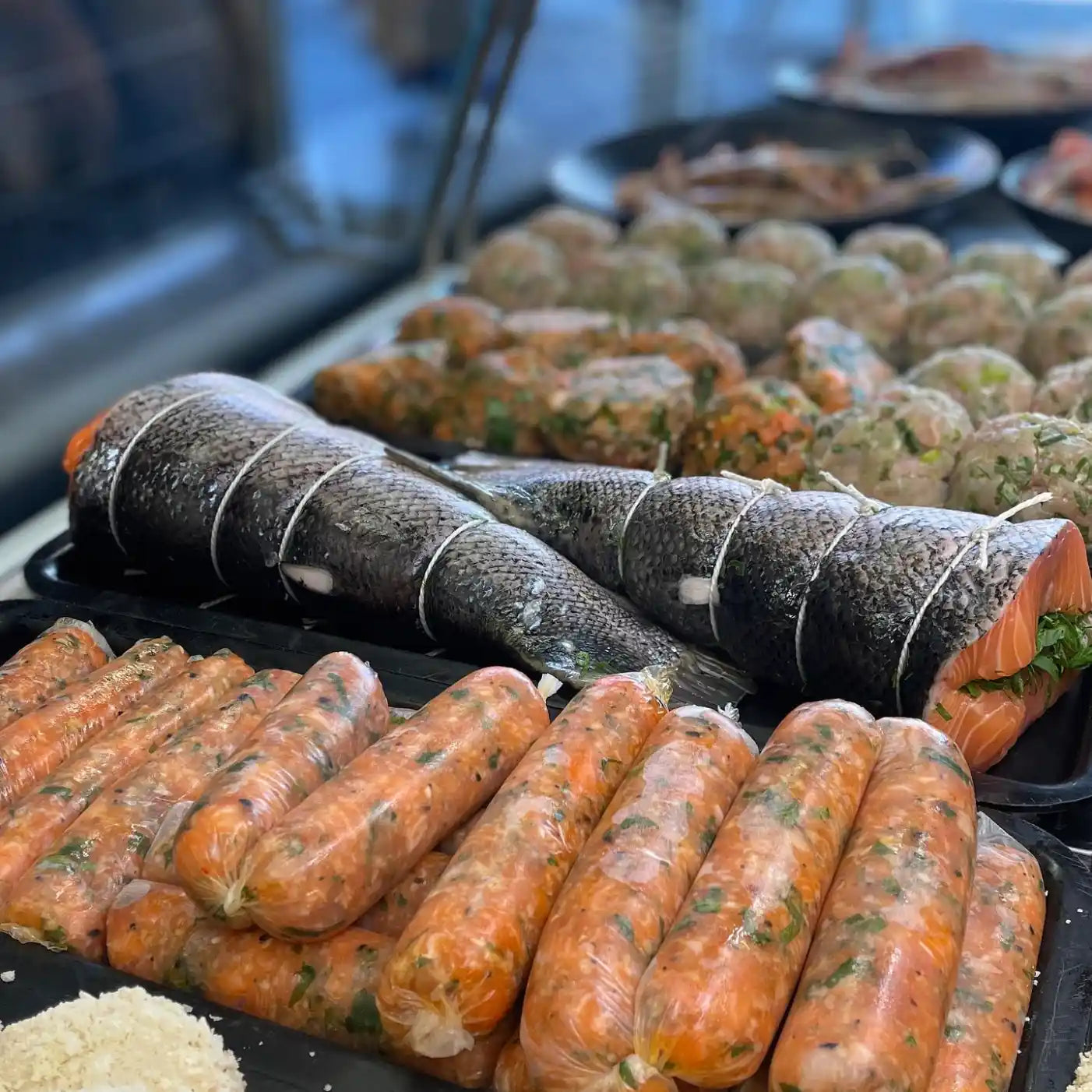 Fresh seafood and meats on a display counter, featuring whole salmon fillets wrapped in string, orange-colored sausages, and round meatballs in the background.
