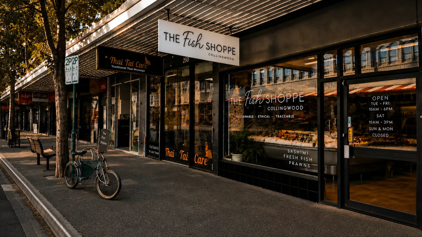 The Fish Shoppe storefront with large glass windows, black tiled facade, awning, and a sidewalk lined with trees on the left.